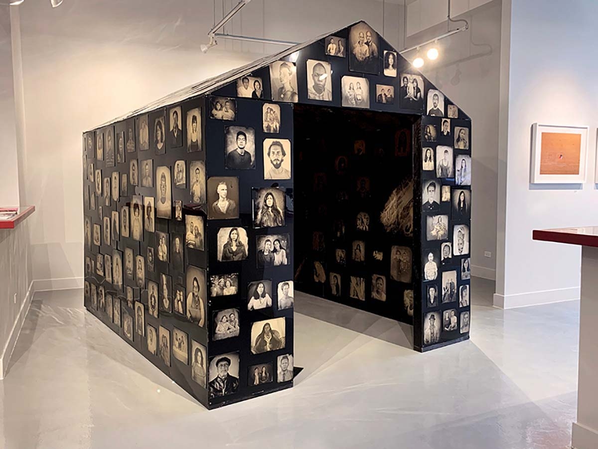 Keliy Anderson-Staley’s “Shelter in Place,” a large constructed house-like building made from blackened aluminum sheathing is covered in 560 tintypes, looms against white gallery walls in this installation view of “What is Home?” at Catherine Edelman Gallery.