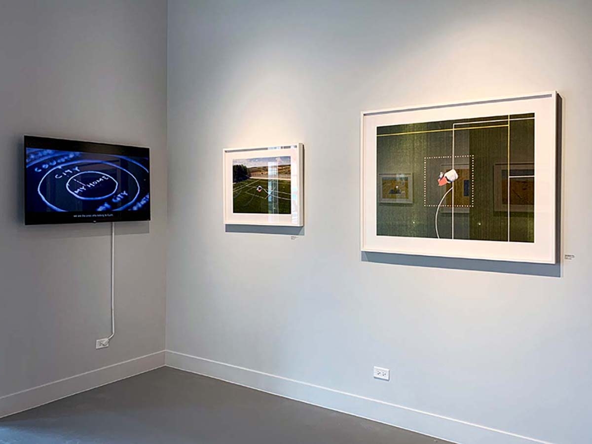 Installation view of What is Home? At Catherine Edelman Gallery. Two framed and mounted photographs adorn the white gallery walls on the right, while a black screen displays a still image from a multimedia work.