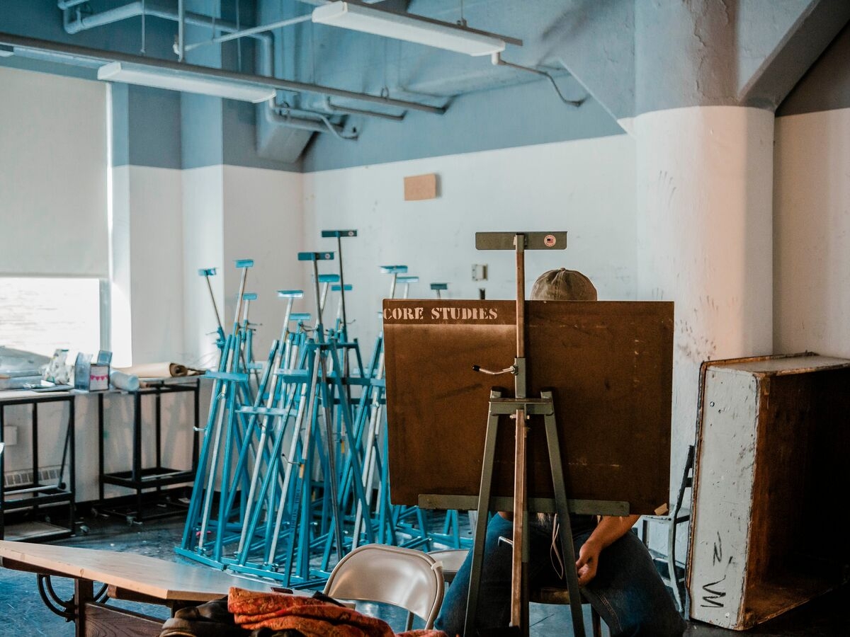 Interior of a drawing studio. In the background is a collection of blue easel stands. A person seated facing the viewer is in the foreground, but is obscured by an easel. white spray paint writing on the easel reads "core studies"