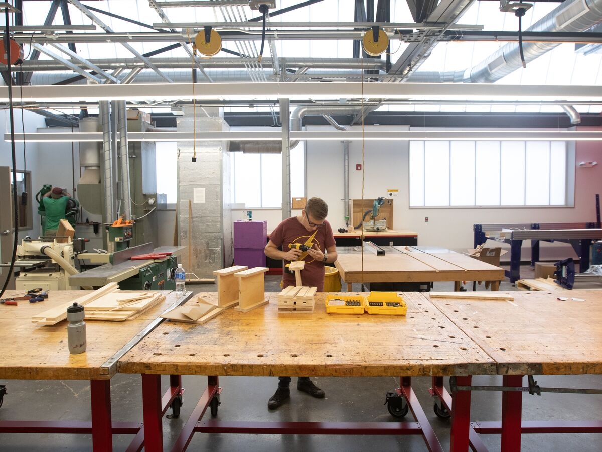 Interior of UArts makerspace classroom. a person with glasses is assembling a wooden object on a large worn wooden table. the room if lofty and bright. 
