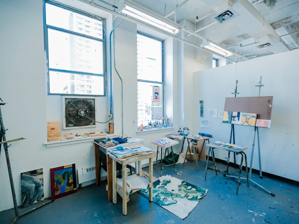 Interior of a painting studio, with various small paintings on easels, tables, and leaned against the wall. a partially painted canvas is on the floor in the middle of the room behind small tables.