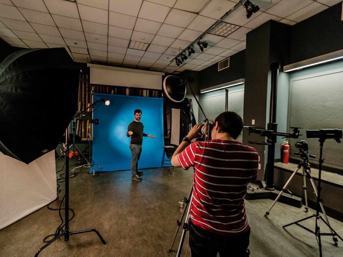 Interior of a photo studio. a person in the foreground is looking through a camera on a tripod at another person standing further back in the room against a blue seamless backdrop. 