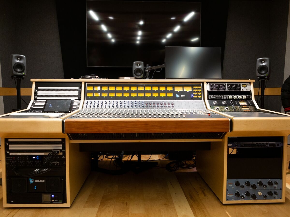 a recording and mixing studio setup, with large mixing console in the center block, patchbays on the left panel and outboard rackmounted equipment on the right. the desk is trimmed in light wood.