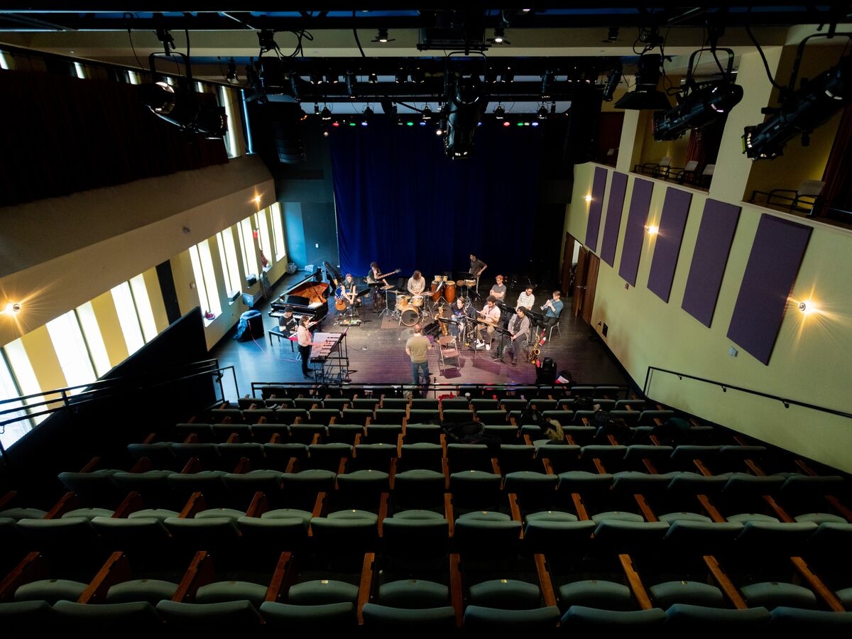 interior of a studio theater space, viewer perspective is from far up in the seating, looking down towards the stage area, where an instrumental ensemble is practicing.  