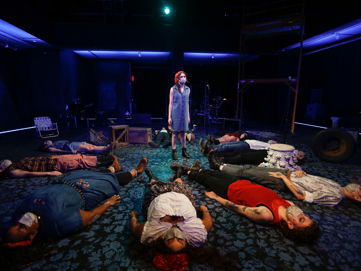 performance of buried. a person in a denim dress and red bandana stands in the middle of a room, surrounded by a circle of people lying flat on their backs. everyone is wearing face masks. the room is illuminated with dim blue lighting.