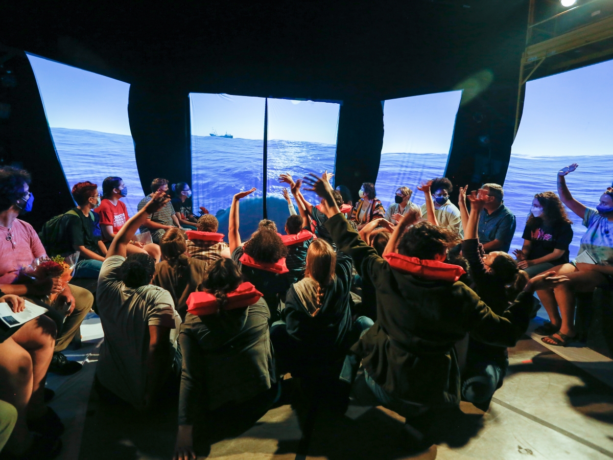stage performance of I want a country. a mass of people in face masks sit in chairs around a group of people seated on the floor. the latter group is wearing red life jackets and many have their hands raised. all people are looking at hanging panels that surround the entire group, displaying an empty ocean in stark sunlight.