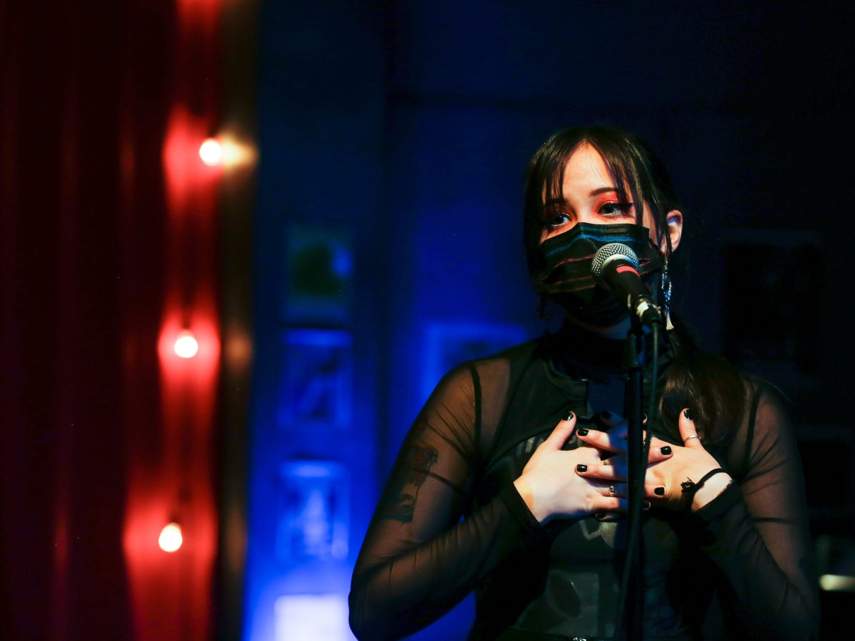 performance of we're gonna die. A person in a black sheer top with bright red eye makeup and wearing a black surgical mask clasps their hands over their chest. their face is close to a microphone. in the background, blurred light bulbs trail vertically along a red curtain, with the remainder of the background washed in blue light.