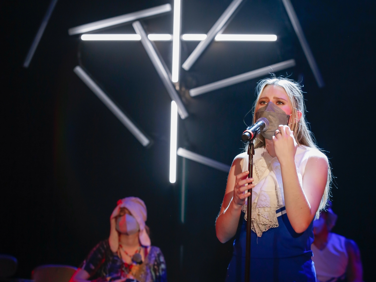 A person in a short blue dress, grey face mask and a frilly ascot sings into a microphone. In the background are two seated masked people, slightly out of focus. on the black wall behind the singer is a jumbled, intersecting collection of tube lights, with several glowing white in a cross pattern.