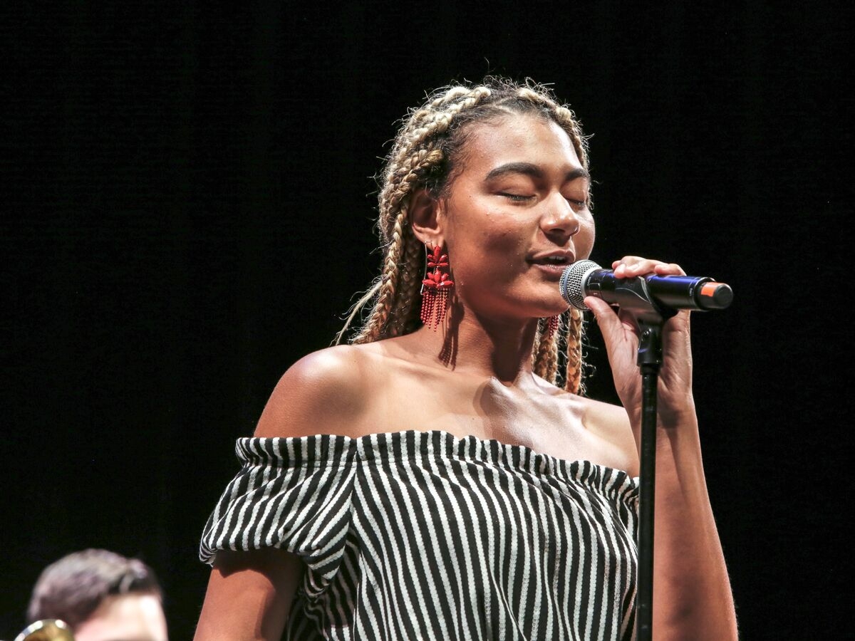 a person with bleached braids wearing a striped top and large dangly red earrings sings into a microphone with eyes closed against a completely black background. 
