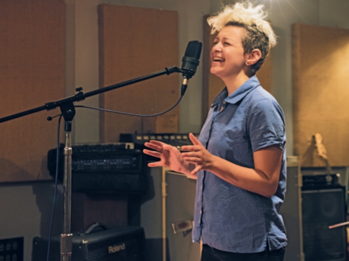 a person with a shock of bleached hear and a blue short sleeved collared shirt sings into a condenser mic in a treated recording studio with amps and recording equipment placed around the room.