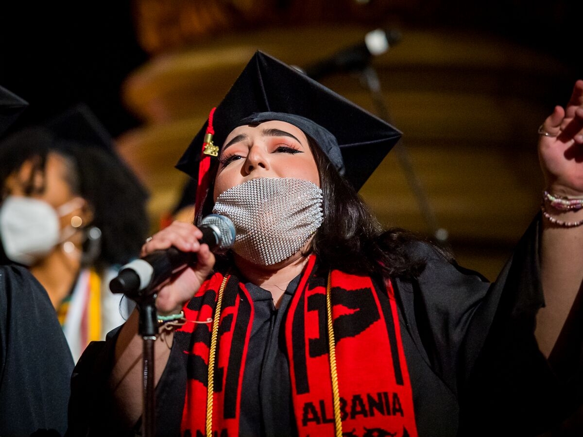 a person in a black graduation robe and cap sings into a mic through a sheer, gem-studded mask. they are wearing a red graduation stole that reads "albania" 
