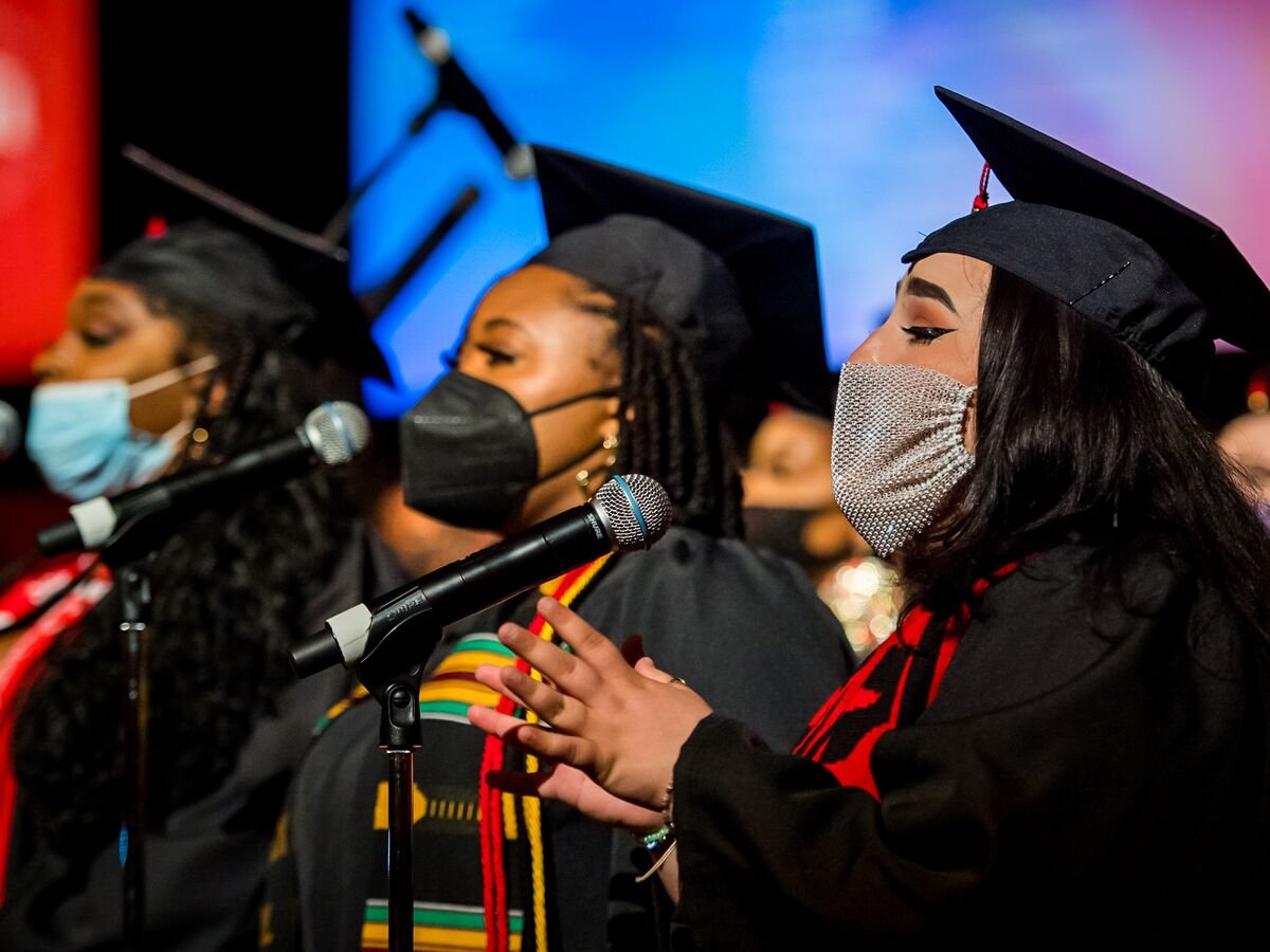 three people in black graduation robes and caps are seen in profile singing into microphones while wearing masks. the person in the middle has a kente-style graduation stole. the person in the foreground (at the right) has a sheer diamond studded mask and has their hands raised. 