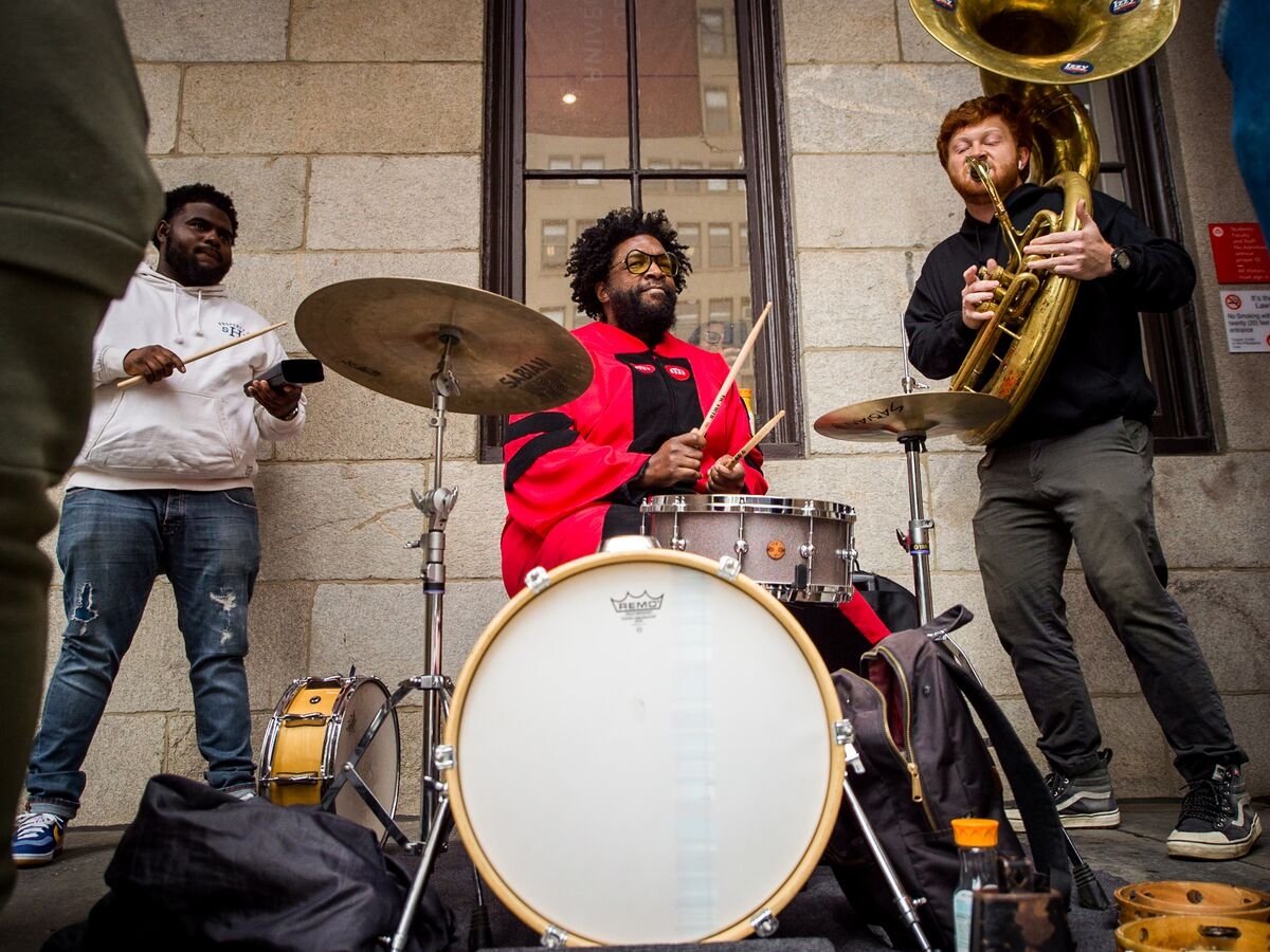 At the center of the image, Questlove is seated behind a drum kit wearing a crimson doctoral robe. Directly to the right, a student in a black shirt plays a tuba, as a student to the left in a white hoodie plays the cowbell. Image perspective from approximately the top of the kick drum. 