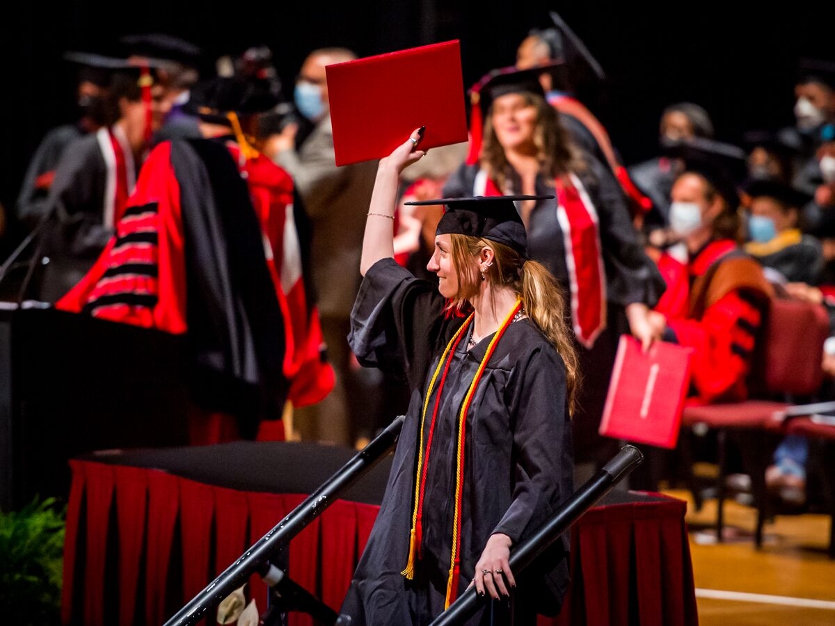 A person in a graduation robe with red and yellow stoles steps down from a stage, bites their lip, and holds a red leather-bound diploma aloft. In the background, people in graduation regalia mingle. 
