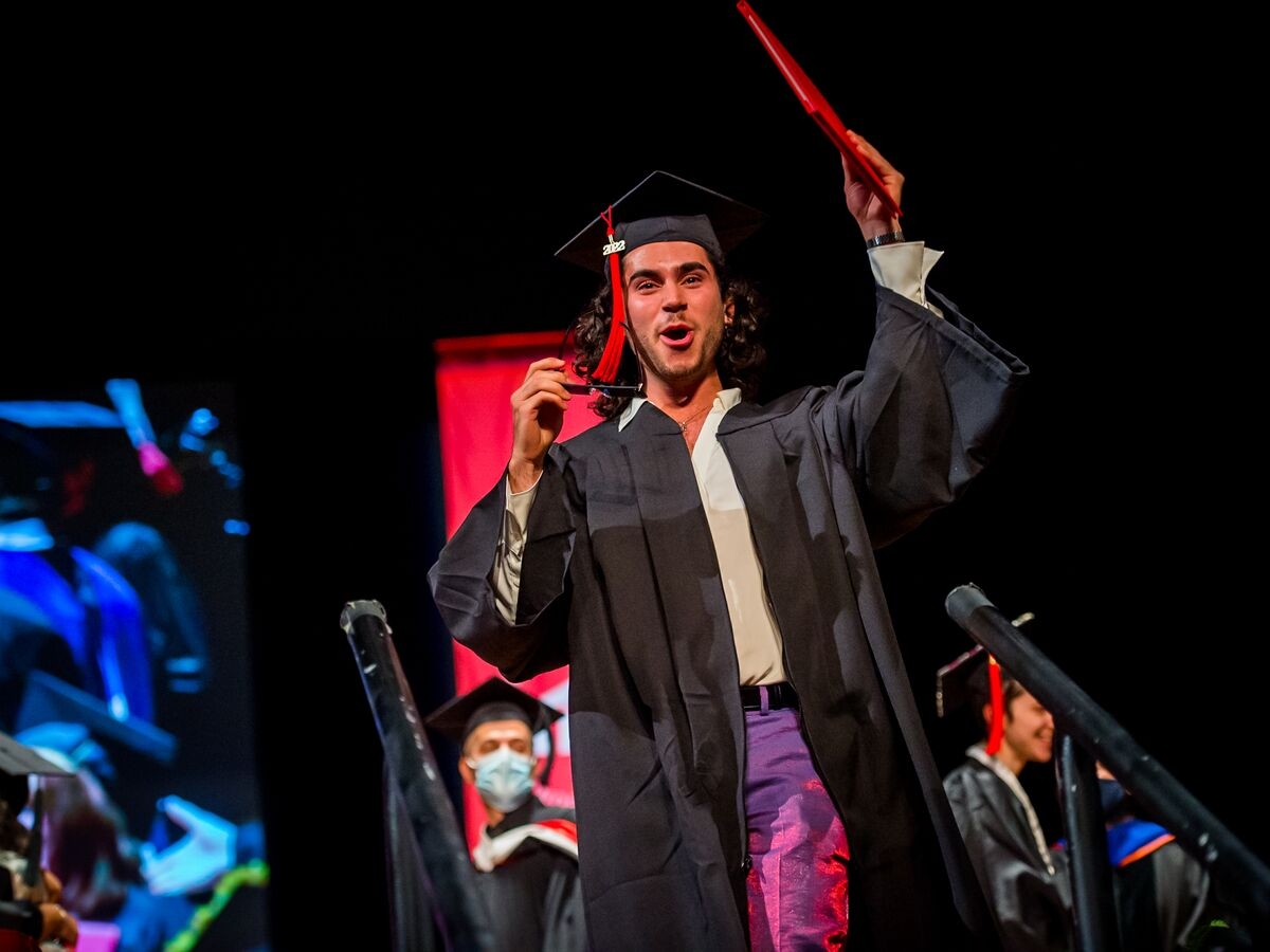 A person in an unzipped graduation robe shouts for joy and holds a diploma of their head while stepping down from a stage. A crimson UArts banner and an azure projection are seen out of focus in the background. 