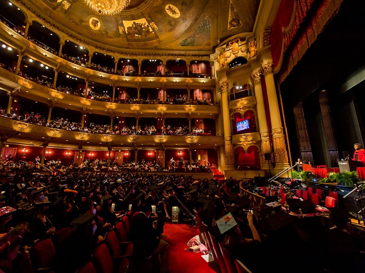 A view across a large, 4-story gilded auditorium that is brimming with people during a graduation ceremony. The ceiling is adorned with gilded paintings and a glowing chandelier, red velvety textures line the seating, floors and walls. A person in a crimson doctoral robe stands on a foliage-adorned stage at the right of the image and addresses the crowd. 