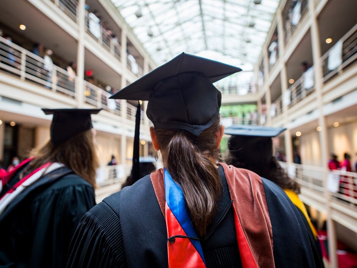 A person in a graduation cap and gown is seen from behind, looking into a brightly illuminated, multi-story indoor courtyard with a glass ceiling. Out-of-focus people can be made out on the three floors of the courtyard behind the railing. 