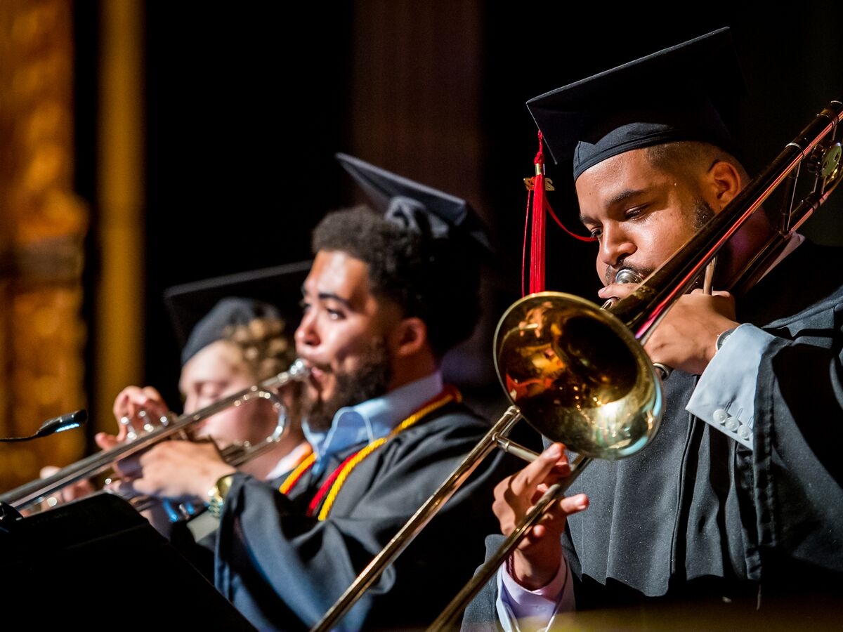 A row of students dressed in black cap and gown graduation regalia over pale blue dress shirts perform on brass instruments. The closest student on the right plays a trombone, the student in the center plays a silver trumpet.
