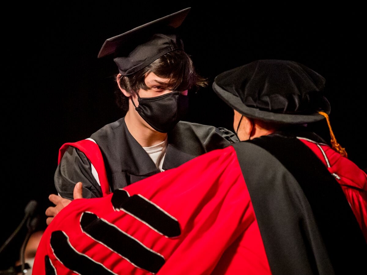 a student in a black graduation cap and gown and wearing a black face mask faces a person in a crimson doctoral robe, seen from the back and over the left shoulder, who is resting their hand on the students arm in a congratulatory gesture. 