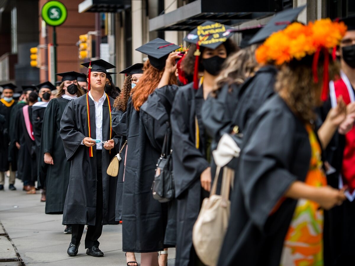 a group of many people dressed in black cap and gown graduation regalia with various flowery and colorful embellishments and sashes stand in a line on the sidewalk. The people in the foreground and background are out of focus, with the image focal point on one person holding a teal surgical mask in their hands and gazing into the distance.