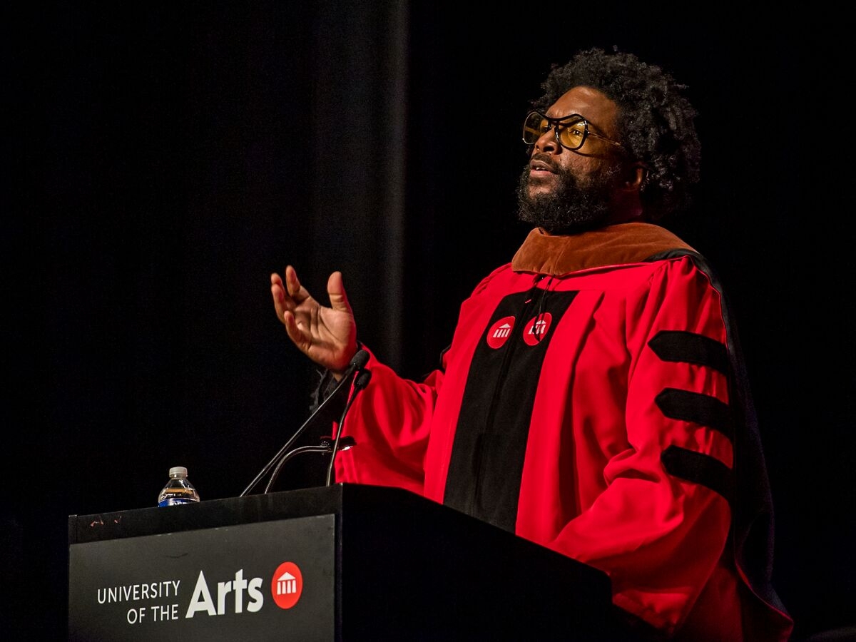 Questlove, seen angled from the right, is dressed in a crimson doctoral robe emblazoned with UArts logos on the chest, raises his right hand and speaks out over two microphones at a podium labeled with a University of the Arts logotype.