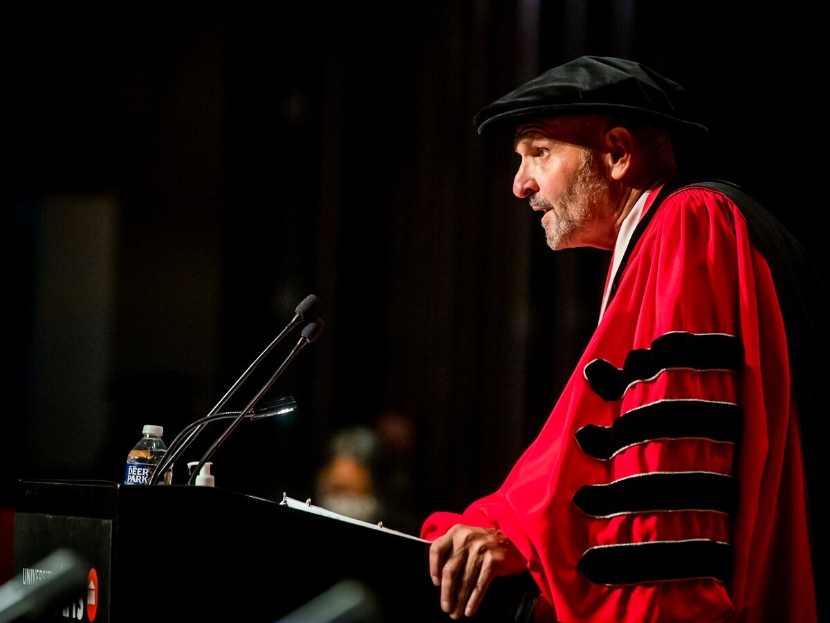 UArts President David Yager, seen squarely from his left, is dressed in a crimson doctoral robe emblazoned with black stripes on the arm, speaks into two microphones at a black podium.