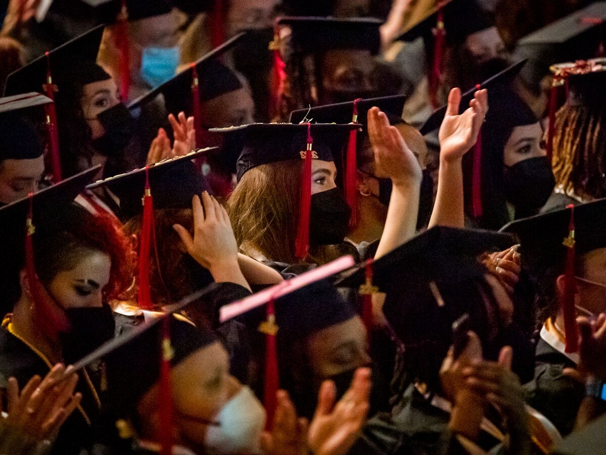 a dense, seated crowd of people in black graduation garb are seen from the side, clapping and looking ahead. Their tassels are red and on the right side of their graduation caps. All are wearing face masks, mostly black ones. The person in the center of the image and most in-focus is holding their hands higher than anyone else around them. 