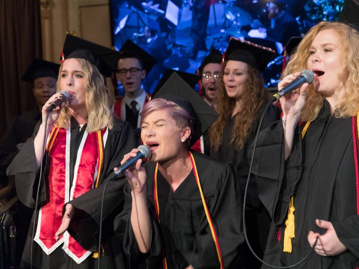 a group of people in graduation caps and gowns all sing together, with three people in the front singing into microphones. the person in the center of the image has their eyes closed, bending down to sing passionately. they have lavender hair and a septum ring. 
