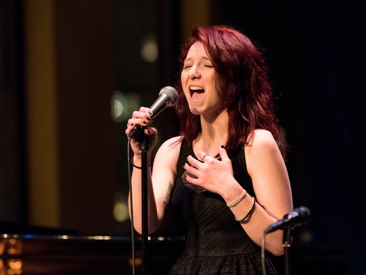 a person with long red hair in a black dress sings into a mic with their eyes closed. the background is out of focus, but reflected stage lights and a possible closed piano lid are implied. 
