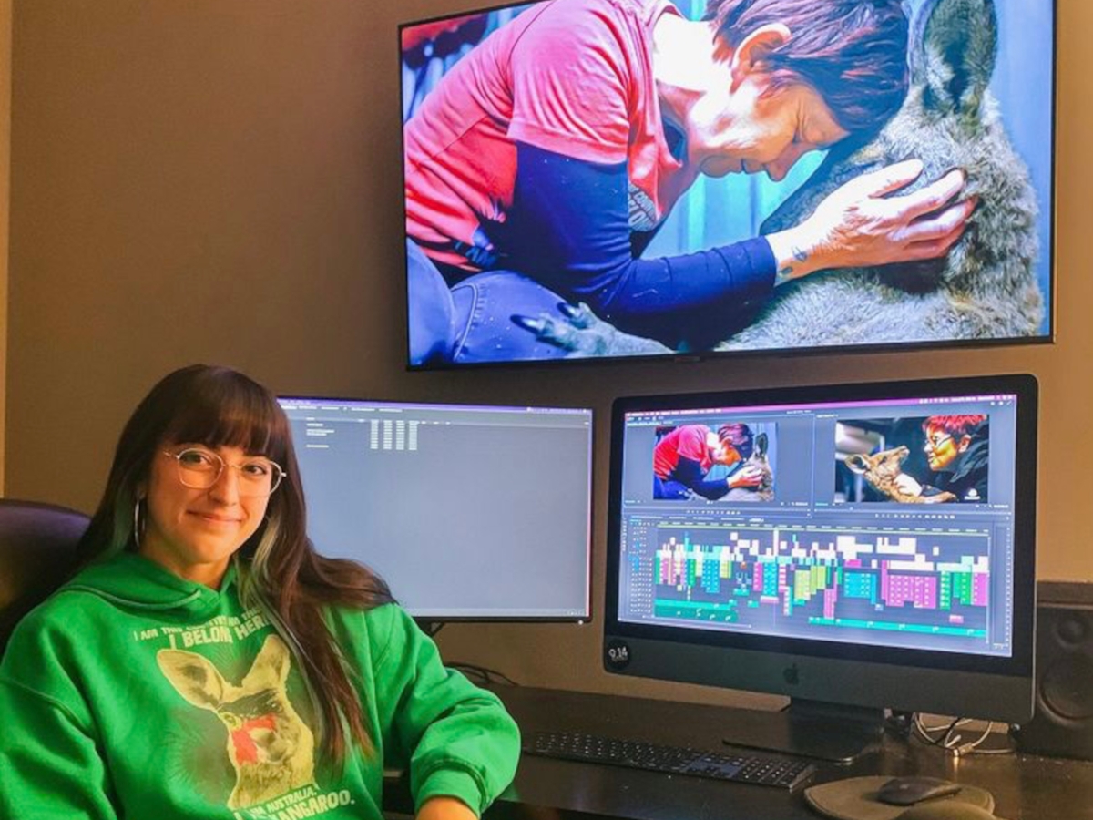 Emily Angelucci sits at a desk with her left elbow on the desk, turned towards the viewer and smiling. she is wearing a green hoodie. on the desk is a double-monitor video editing setup, showing a detailed project being cut. 