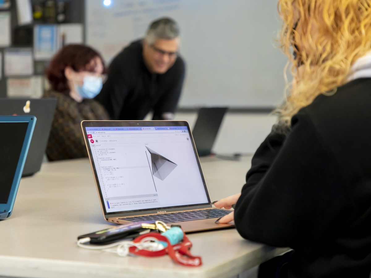 A student is using a laptop in a classroom. Another student is shown in the background, blurred, and Juan Parada, IxD program director, is leaning over their laptop. 