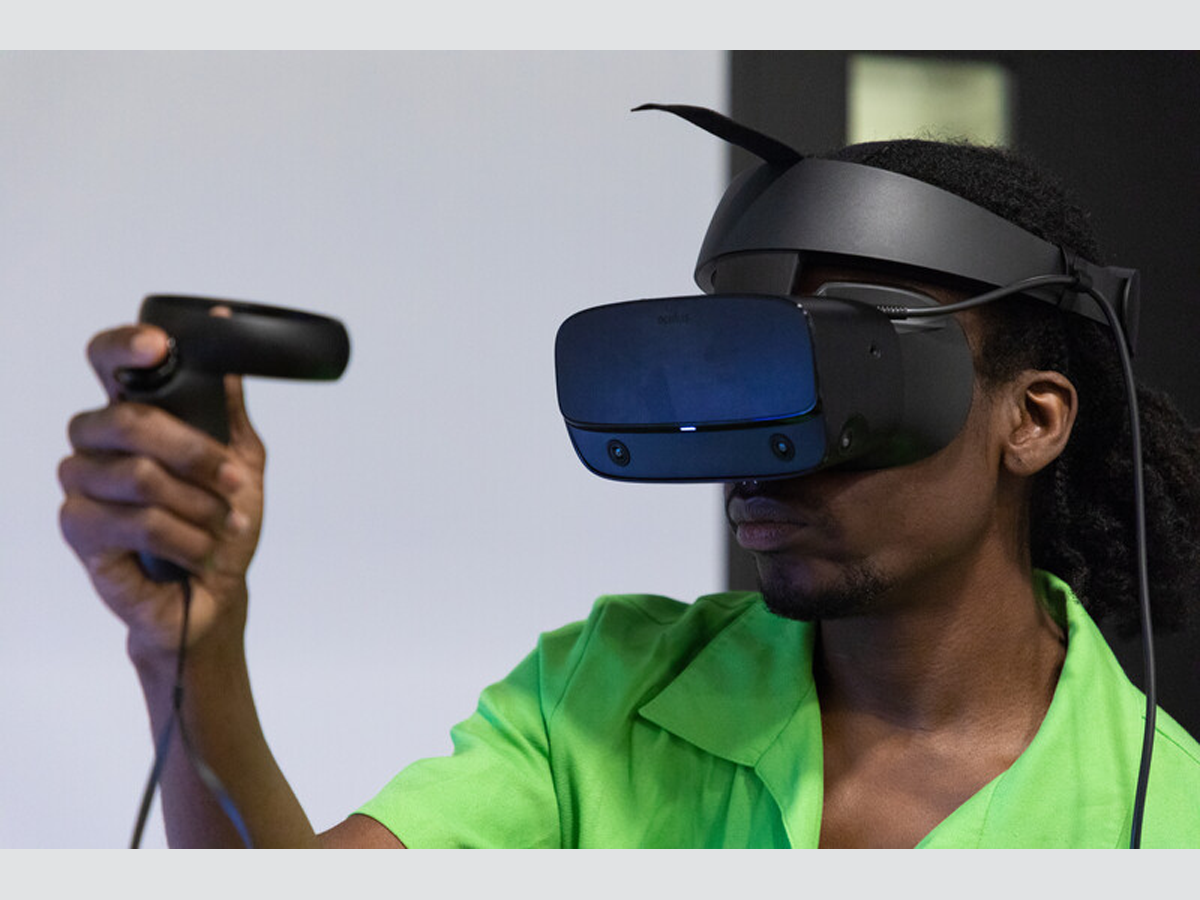 A person with dark skin wearing a lime green shirt is wearing VR goggles and holding a VR controller in their hand.