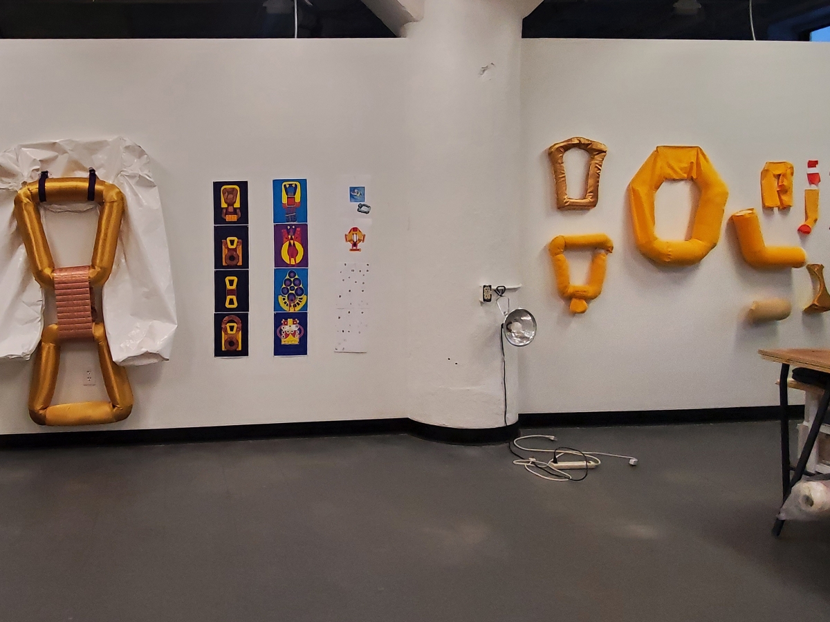 Interior of an art studio. Across from the viewer is white drywall, bisected by a large white concrete pillar. The right half of the studio wall displays fabric loops and shapes, in gold hues as well as lifevest-yellow. The left half of the studio wall are white fabric pants-like U shapes, one of which is draped over a golden figure-eight-type object. Between this object and the pillar are a variety of small prints, each with images resembling the various compositions and objects on the walls. 