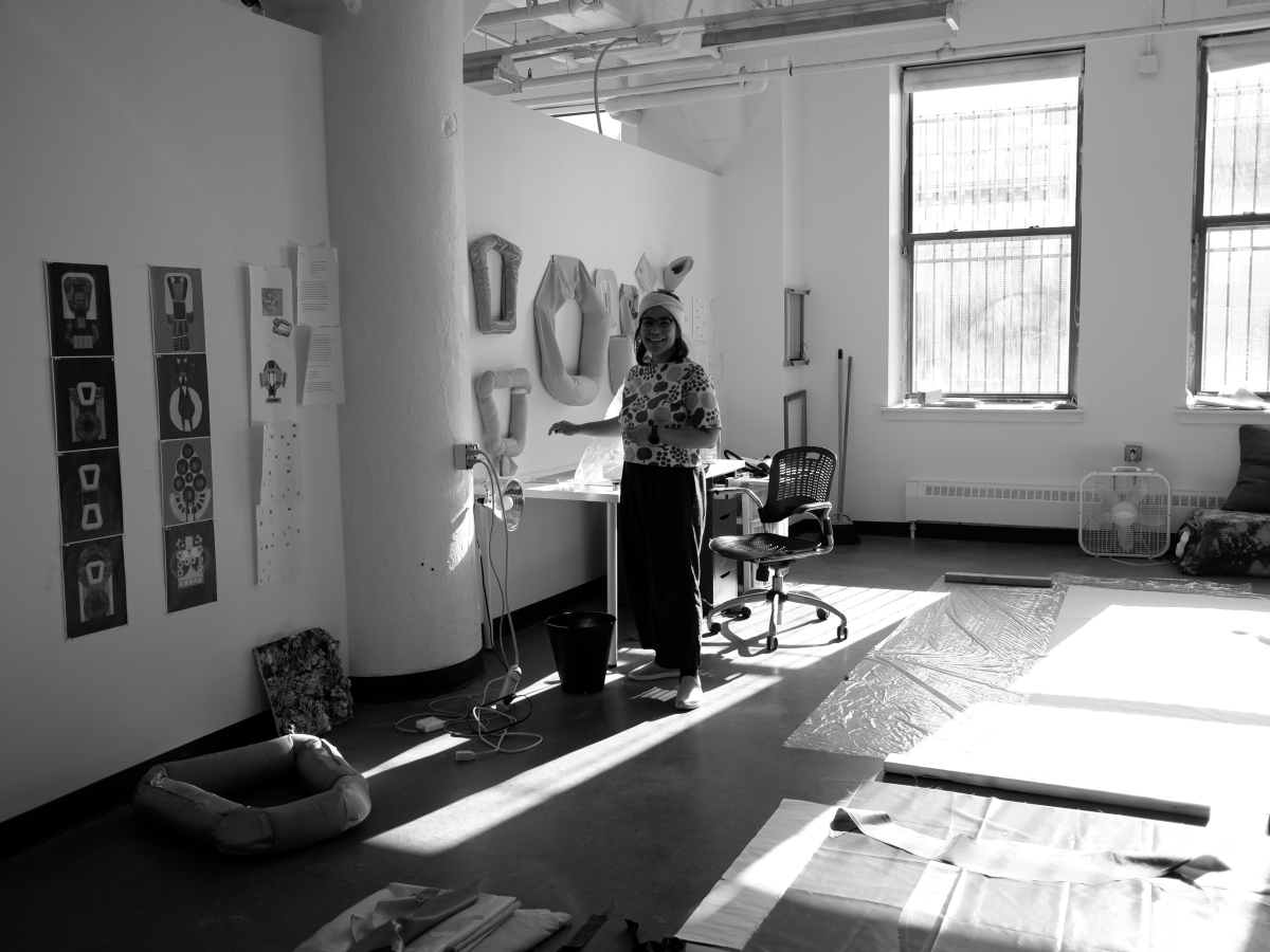 A person stands next to a desk in a large, open studio as sunlight streams in. the left wall is white drywall covered in artworks, interrupted by a concrete pillar. The right, back wall has large windows that allow large beams of light to fall across the floor, which is covered in textiles and plastic sheeting. The person is wearing a floral shirt and is smiling. Image is black and white.