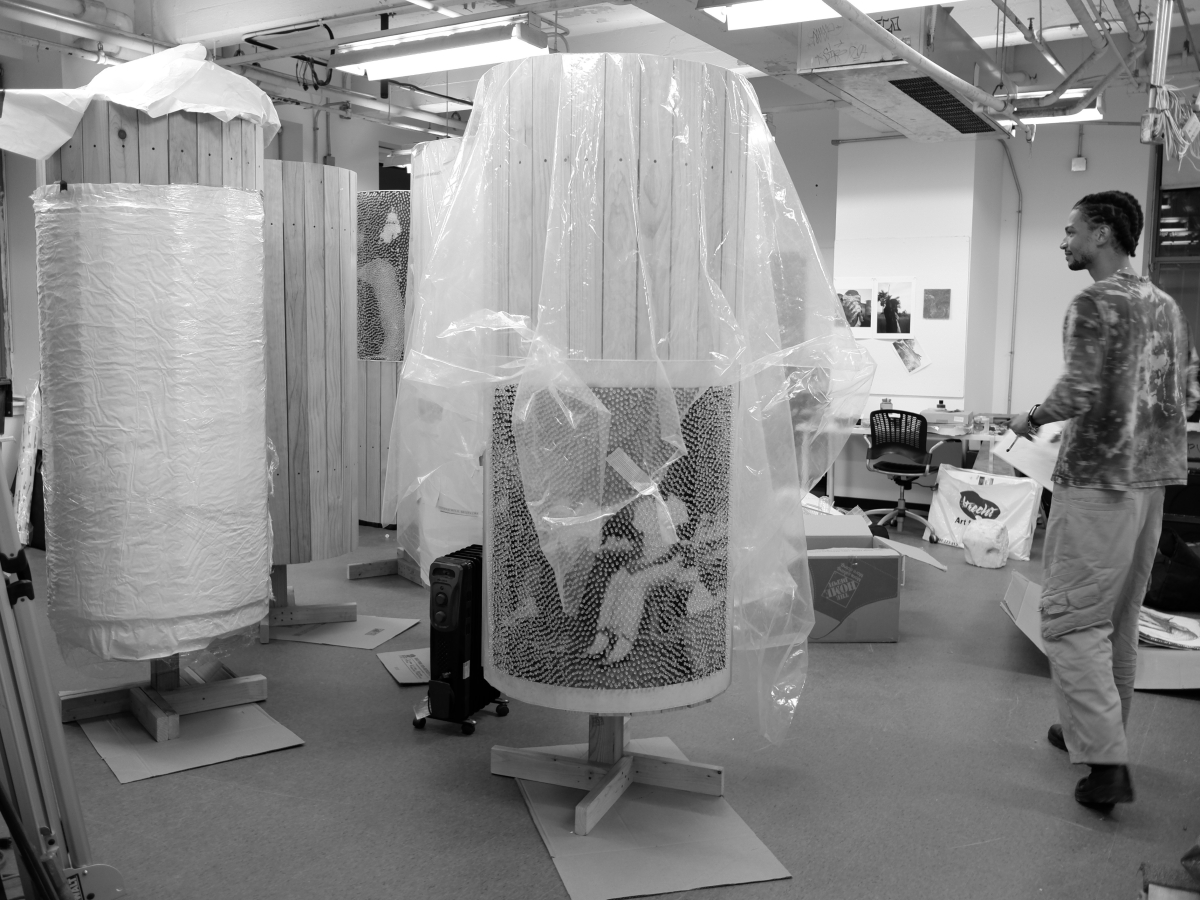 A large studio space containing several large, wooden cylindrical artworks, variously draped or wrapped with plastic. The wooden pillars are free-standing on small wooden bases. The central pillar has a pointillist, mosaic-like image wrapped around its wooden boards. A person in a bleach-dyed shirt walks around the pillar collection on the right of the black and white image of the studio. 