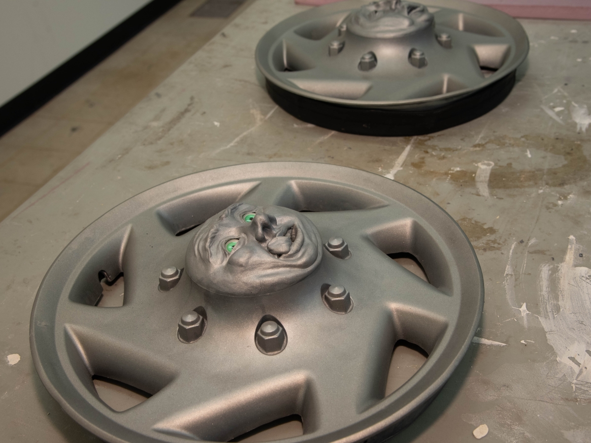 Artworks laid on the floor of a studio. These are two stylized hubcaps, each in a generic standard silver with a slanted seven-spoked star-like design. However, the center of each hubcap contains a raised face. The closer one has a slightly upturned, big nose with large browse, bright green eyes, and a tongue sticking up across its lip. The further hubcap’s face looks like a droopy, tired, slack-jawed expression, though it is not clearly visible in perspective. 