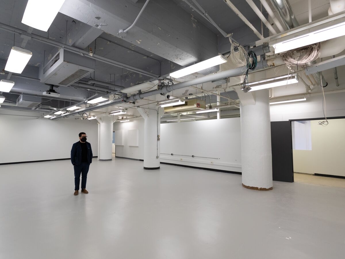 Interior of new studio space shortly after construction in Anderson Hall. It’s a stark utilitarian space with a high, open ceiling with pipes, wires, ventilation, and fluorescent lights. The walls are bright white, as are three support pillars. There is a black door at the far right of the photo. The base of the wall has a black trim along the floor. A person wearing an all-black outfit and black face mask stands below the lights, looking towards the right.  