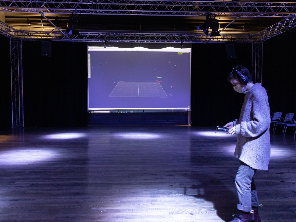 An empty room rimmed with black curtains and supporting lighting trusses along the ceiling. at the rear wall of the room is a projector screen showing mapping software, with a coordinate grid mimicking the room. In the foreground is a person wearing a facemask and headphones holding a tablet, seen in profile. The room is illuminated with pale lavender-hued spotlights pointed down and warm lights towards the ceiling. 