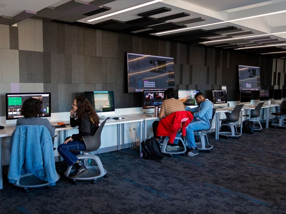 A long room with walls covered in slate gray acoustic paneling and a row of desks against the wall. The white desks each have computer screens on them, displaying video editing software. People sit on rolling chairs at the workstations, looking at project timelines or glancing at notes. Away from the wall, the room is empty, with the floor covered in splotchy gray carpet. 