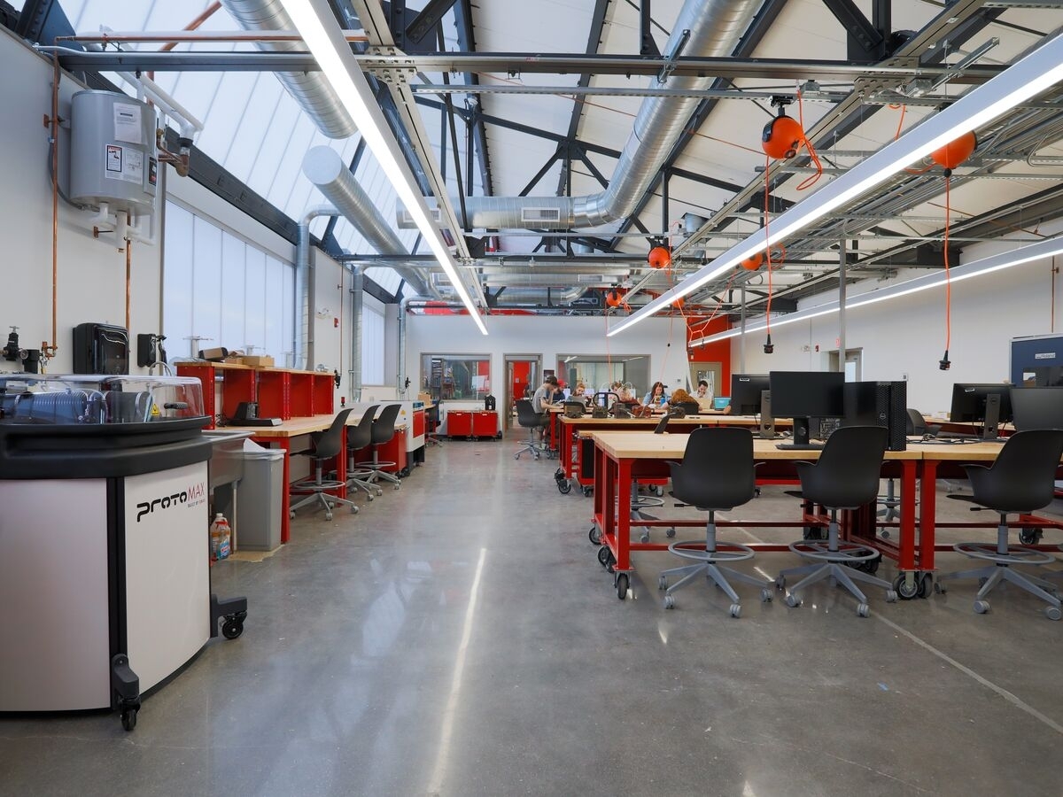 Interior of the UArts Makerspace: an industrial-type space with a very high, slanted ceiling and windows for additional illumination. Below the ceiling, which slopes downwards to the right, are ventilation pipes and long fluorescent lights. The floor is shiny and buffed, and throughout the space are wood workshop tables with red frames on casters. 