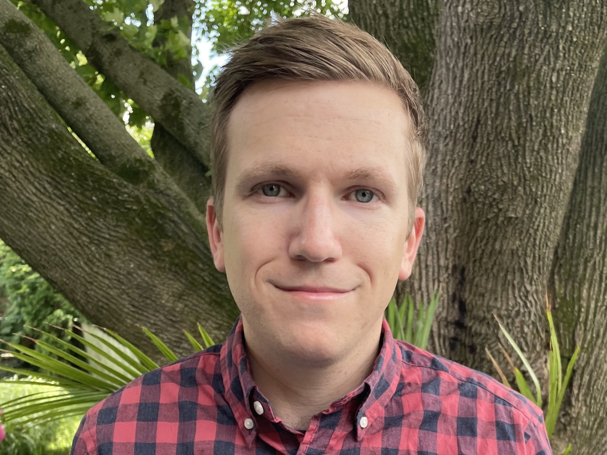 headshot of Timothy Potts, pictured wearing a black and red checkered collared shirt against a background of a tree in a lush garden. timothy is smiling, and has dark blond hair swept to the side. 