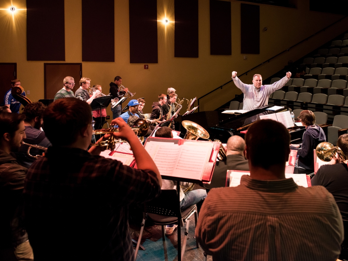 a dynamic photo of a rehearsal at caplan recital hall, seen from stage right behind trumpet players in a large band of instrumentalists with other brass instruments. towards the right where the green seating becomes visible is a large grand piano with a conductor in a white button-up shirt with both hands held aloft in fists. 