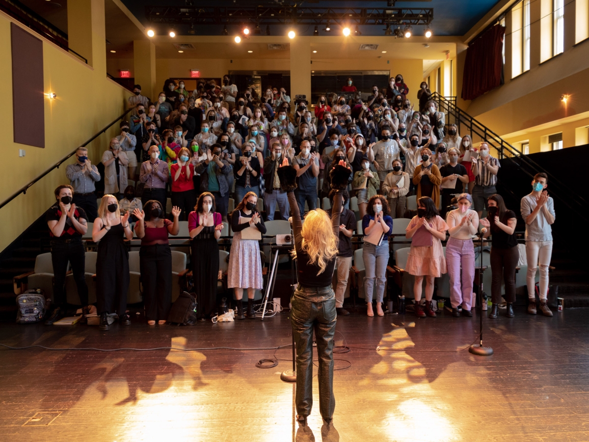 a view of caplan recital hall from the stage. a person with long blond hair and leather pants faces the audience with both hands in the air. the rakes auditorium seating is packed with people who are all standing and interacting with their hands, either clapping or raising them. people are wearing face masks. 