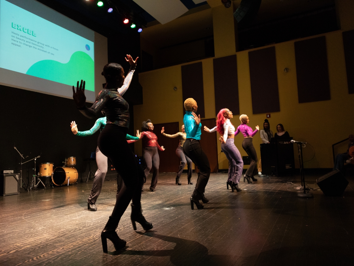 a group of dancers in brightly colored tops and tight pants are dancing on large high heels on the floor of caplan recital hall. the view is from stage right looking towards the dancers in low lighting, with acoustic paneling seen against the yellowish rear wall and a drum kit atthe back of the stage on the left. a projection above the event on the image's left reads "excel" in green blocky type. 