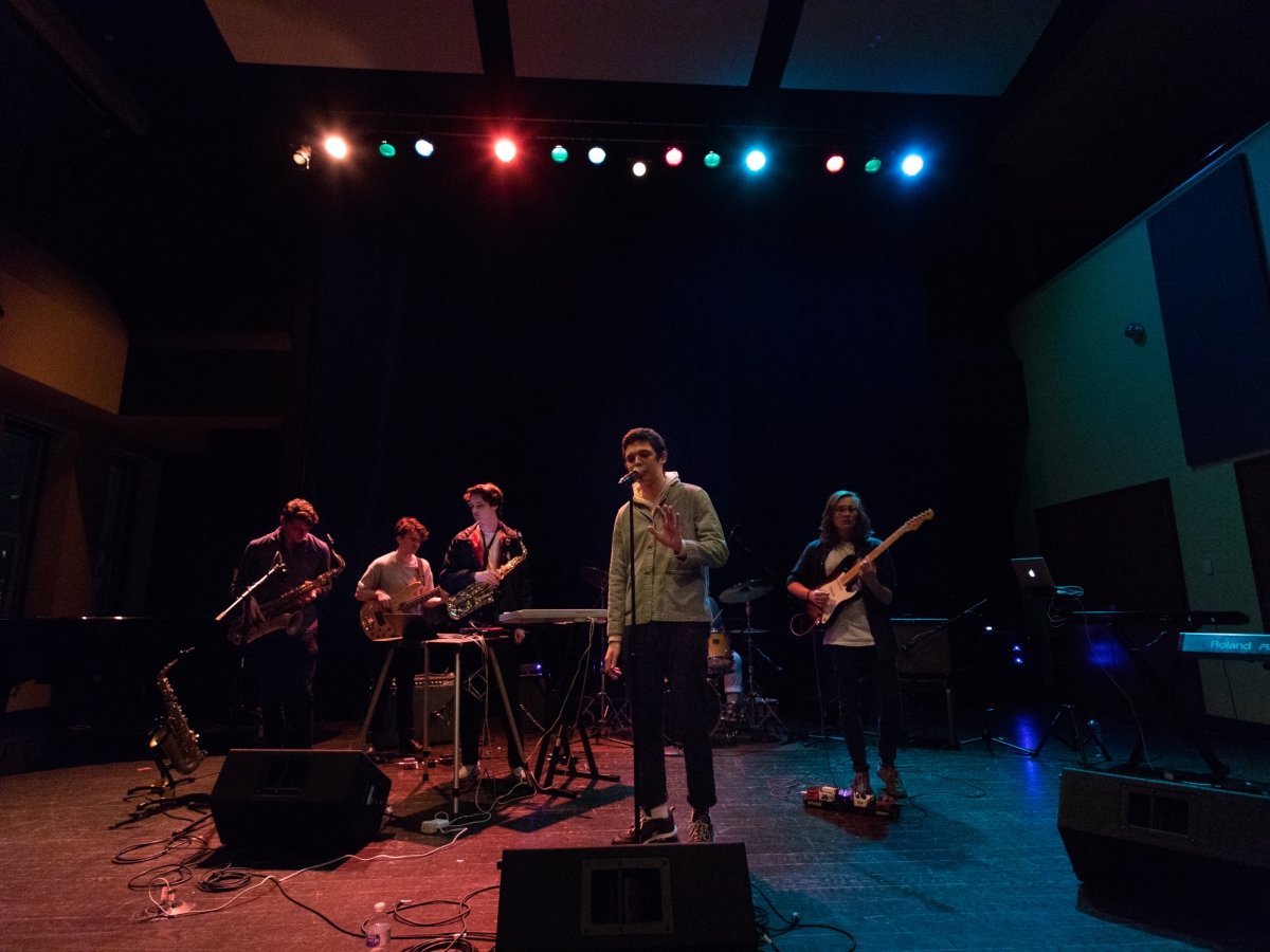 a five person band performs under low, moody light on the floor of the caplan recital hall. stage lights overhead wash the band in red and blue. a person in the center sings into a tall microphone. 