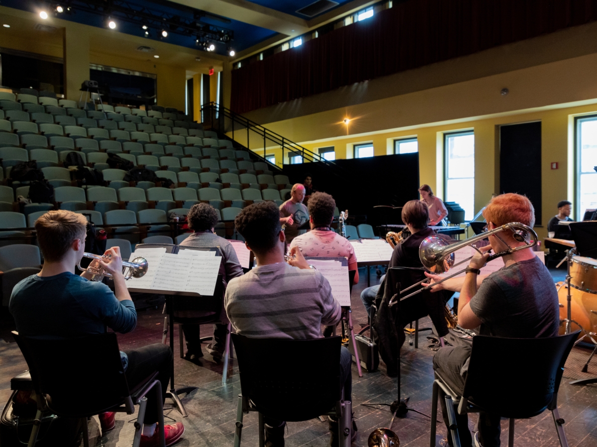 a band rehearses in caplan recital hall. the view is from stage left looking over the shoulders of seated band members with brass instruments facing the conductor. 