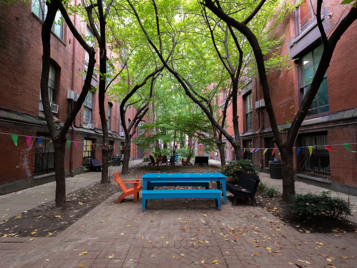 a view of furness courtyard from within, looking out towards 15th street. in the foreground is a large blue plastic picnic table with matching benches and an orange plastic lawn chair. twisting trees ring the seating areas, themselves ensconced by the dark red brick walls of the building. 