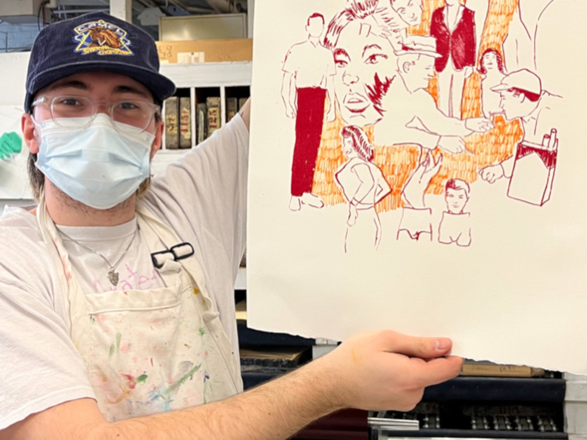 a person in clear acrylic glasses, cap, mask, and stained apron holds aloft a lithograph print with sketched our characters in red with an orange filler texture. 