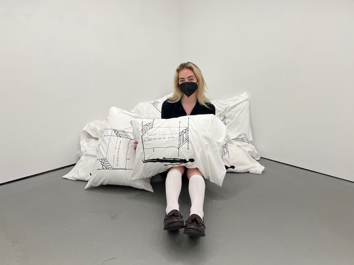 a person in knee high socks and loagers and a black facemask with long blond hair sits in a studio space on a pile of pillowcases with screenrptings of a bed with a shadowy figure