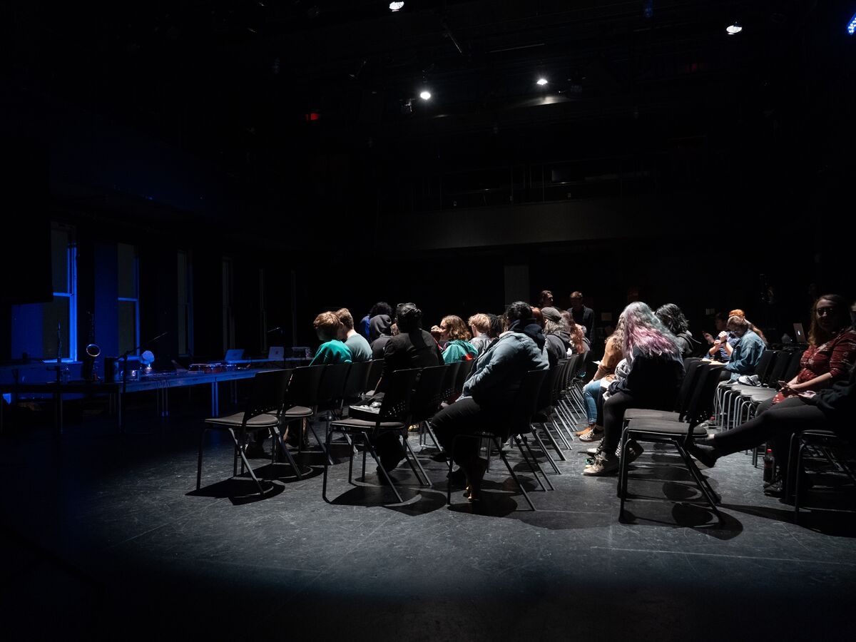 an image of the seating during the Out Of The Box performance in the caplan studio theater. the seating is seen in profile from ithe left of the audience, and the chairs are all on the flat floor - not raked. the audience is illuminated with white light while the empty stage is washed in blue light. 